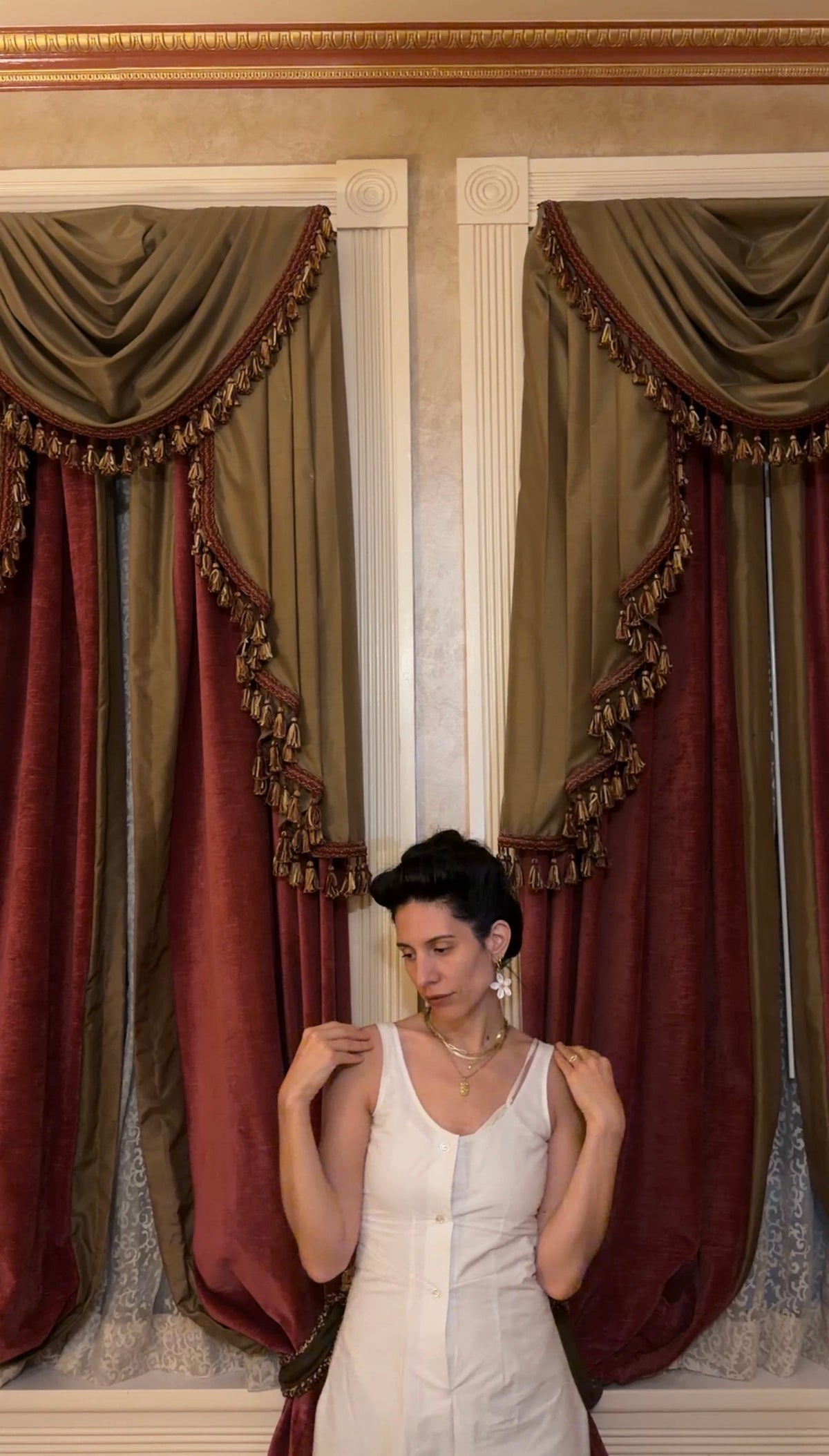 A woman wearing a white sleeveless romper stands in front of ornate red and olive drapes with gold fringe trim in an indoor room with classical columns.