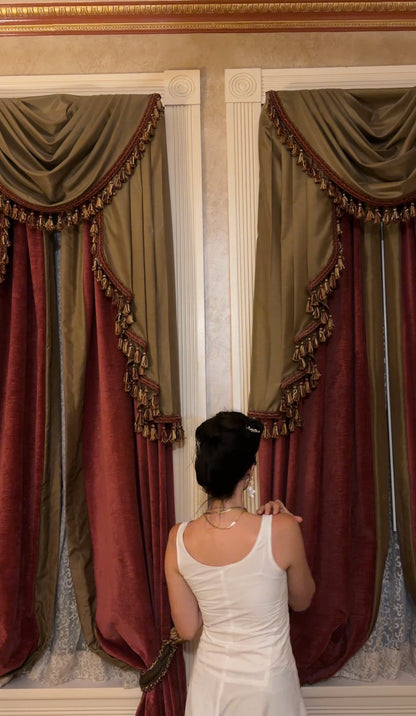 A woman in a white romper stands with her back to the camera, looking at ornate drapery curtains in a classic room. The curtains feature olive green and deep burgundy velvet panels with gold fringe and valance details.