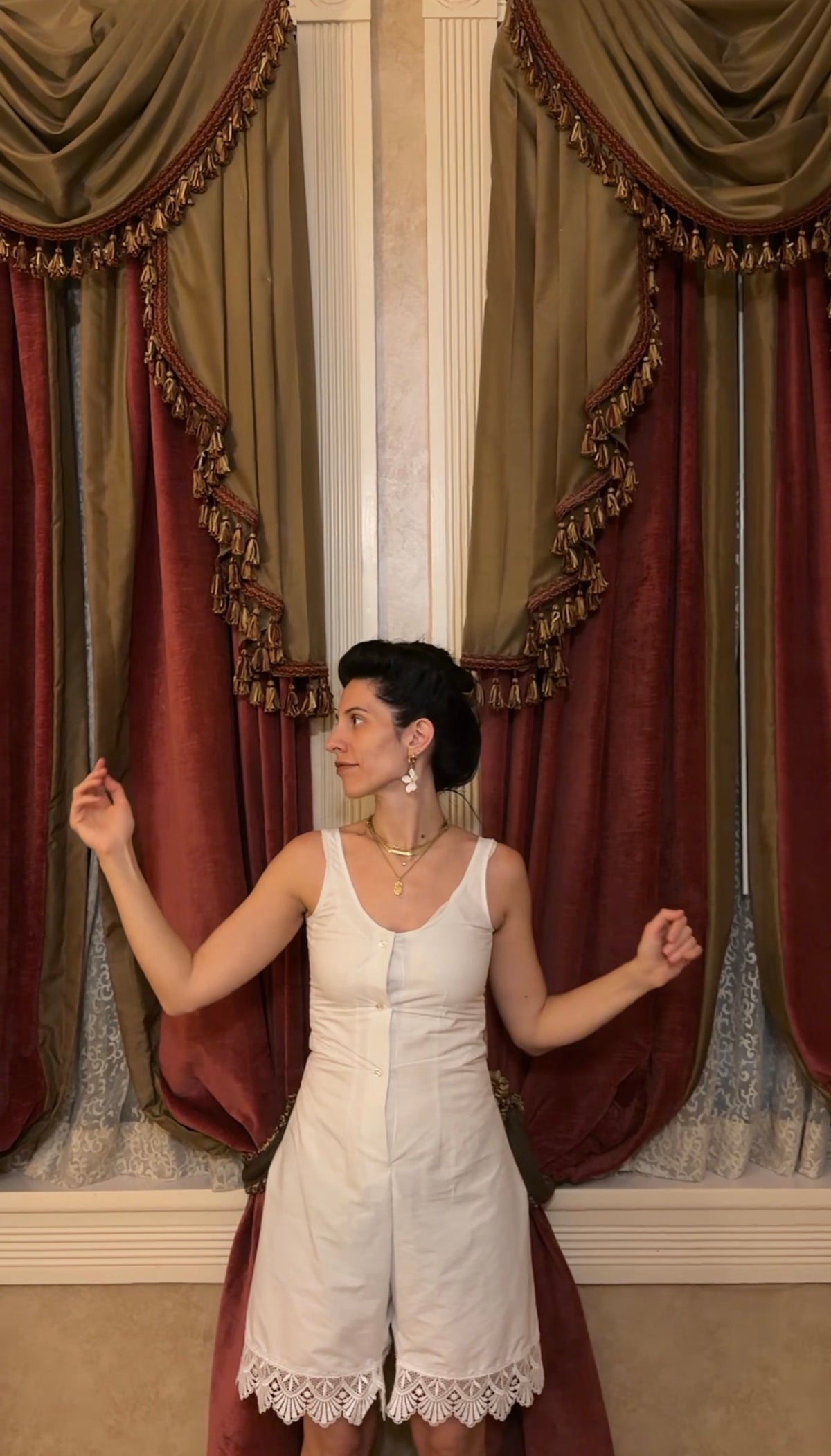 A woman wearing a white sleeveless romper with lace trim stands in front of ornate burgundy and gold curtains.