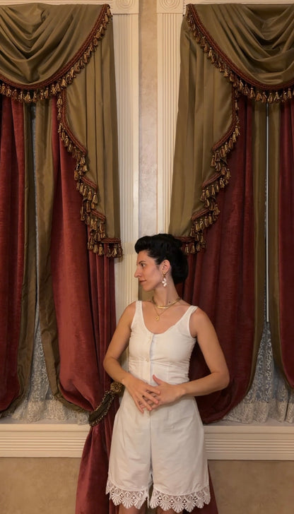 A woman stands indoors in front of ornate drapes, wearing a white sleeveless romper with lace trim at the hem. The setting features rich burgundy and olive curtains with gold fringe detailing.