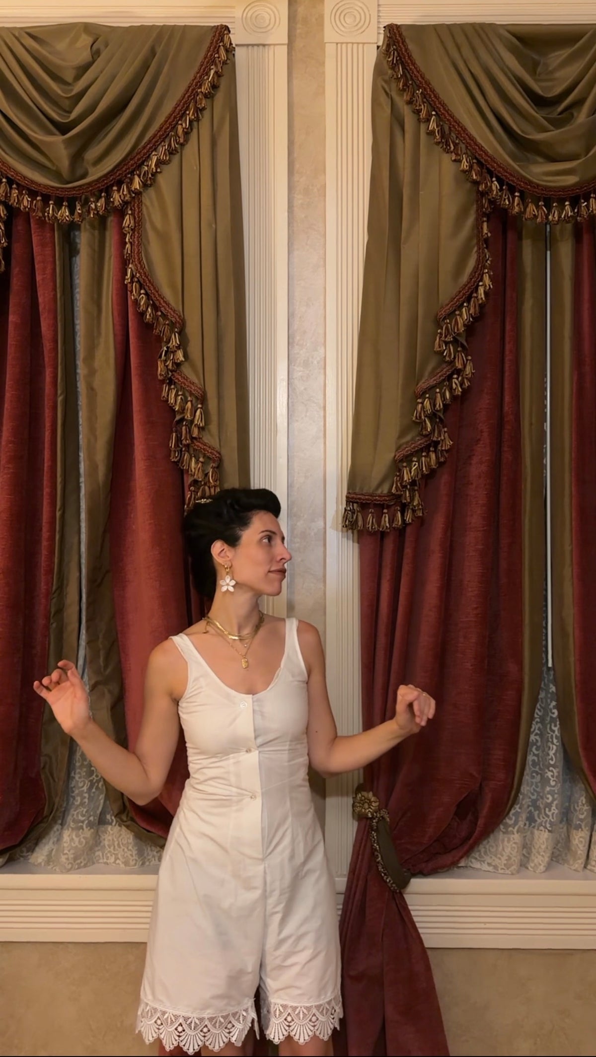 A woman wearing a white sleeveless romper stands in front of ornate drapes with olive green and burgundy velvet fabric and gold fringe trim.