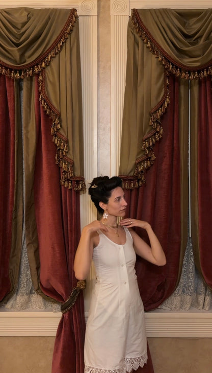 A woman wearing a white sleeveless dress stands in front of ornate drapes with olive green and burgundy velvet panels and gold fringe trim.