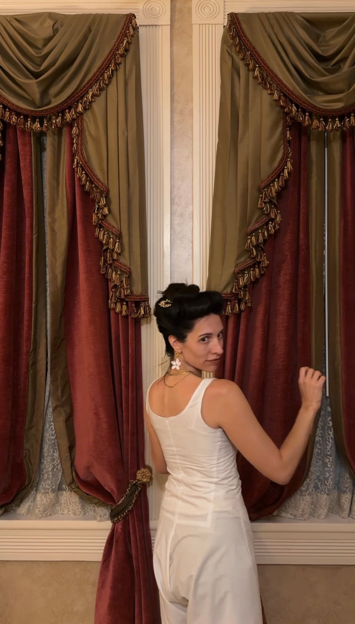 A woman in a white sleeveless jumpsuit stands in front of ornate red and olive drapes with gold fringe, looking back over her shoulder.