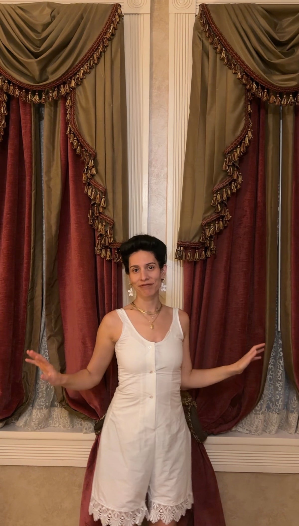 A woman wearing a white sleeveless romper stands in front of ornate red and gold curtains, gesturing with her hands. The romper has a scoop neckline, button-front closure, and a lace-trimmed hem.