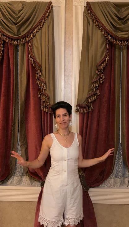 A woman wearing a white sleeveless romper stands in front of ornate red and gold curtains, gesturing with her hands. The romper has a scoop neckline, button-front closure, and a lace-trimmed hem.