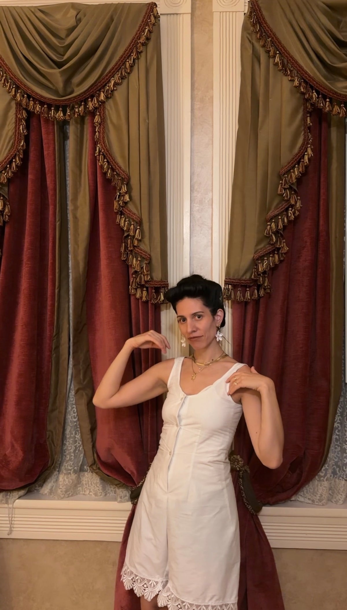 A woman modeling a white sleeveless dress with lace trim, standing in front of ornate burgundy and olive drapes with gold fringe.