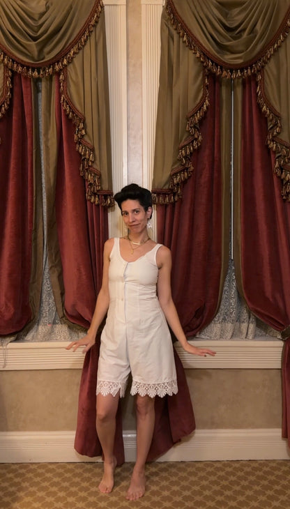 A woman stands barefoot in front of ornate burgundy and gold curtains, wearing a white sleeveless romper with lace trim at the hem.