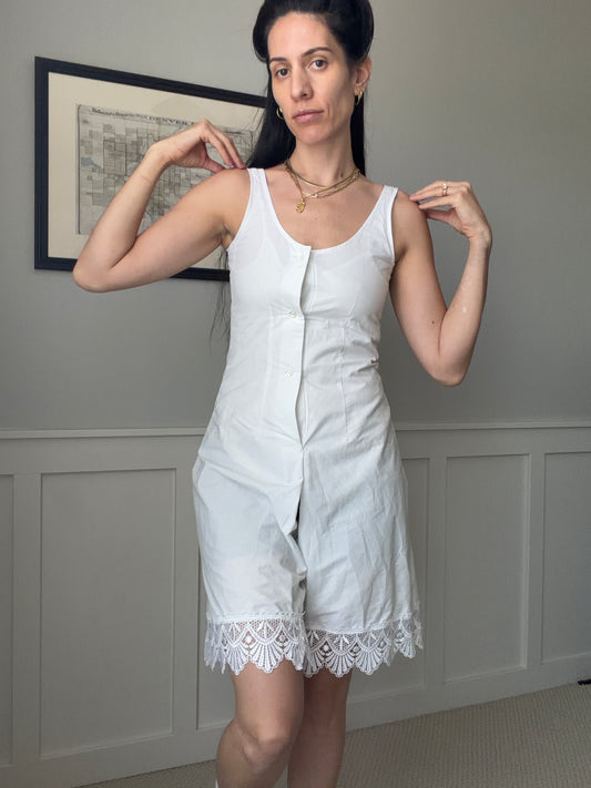 A woman modeling a white sleeveless linen romper with a button-front closure and a white lace hem, standing in a room with gray paneled walls.